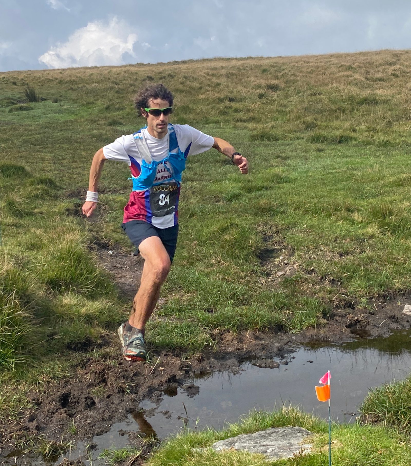 A runner wearing a blue and white racing outfit with bib number 84 jumps over a small muddy patch on a grassy trail. The sky is partly cloudy, and there is a small orange flag marking the trail ahead.