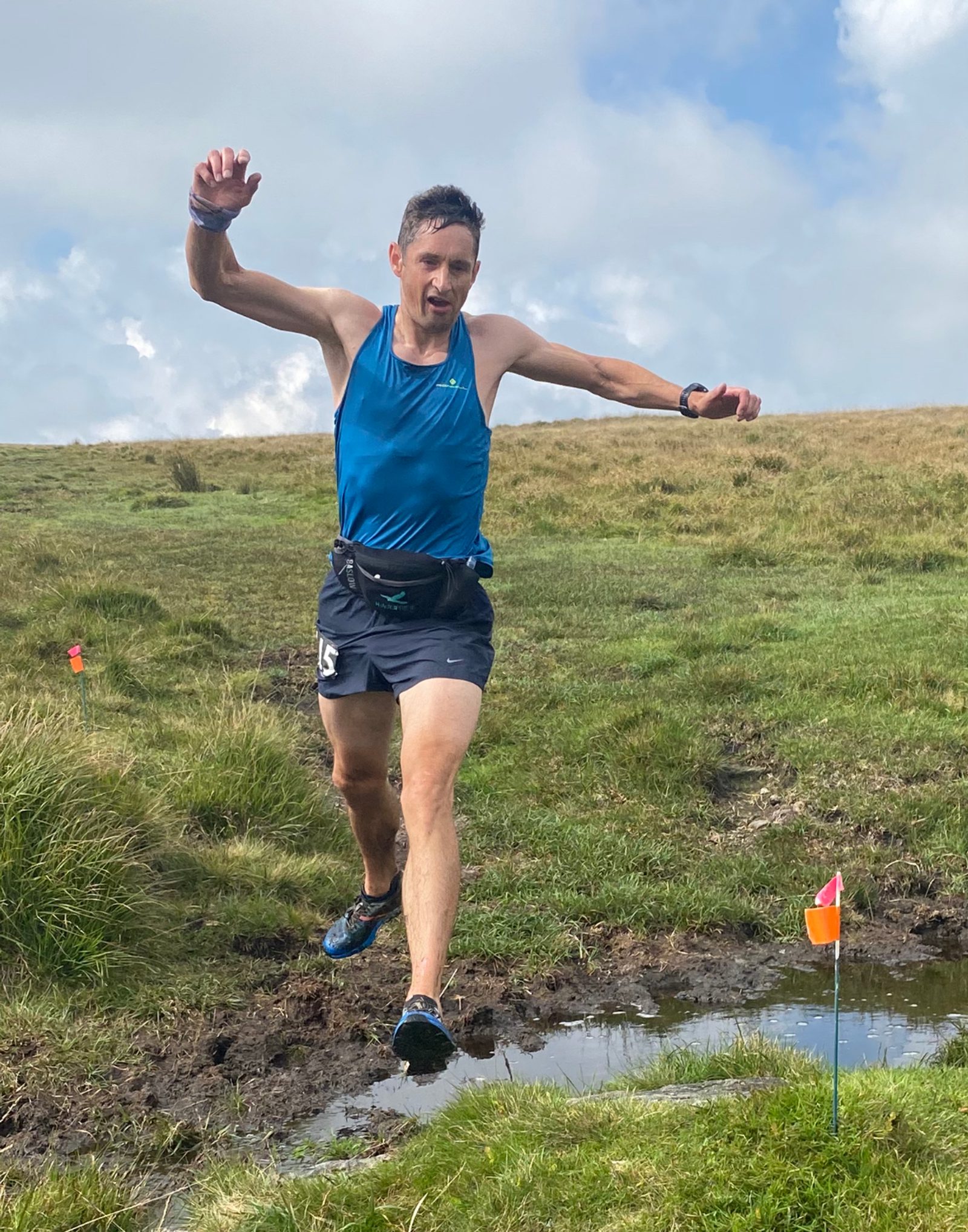 A man in a blue tank top and black shorts is mid-leap over a small muddy puddle on a grassy hillside. He appears focused, with arms outstretched for balance. A small red and white flag marker is visible to the right of him. The sky is partly cloudy.