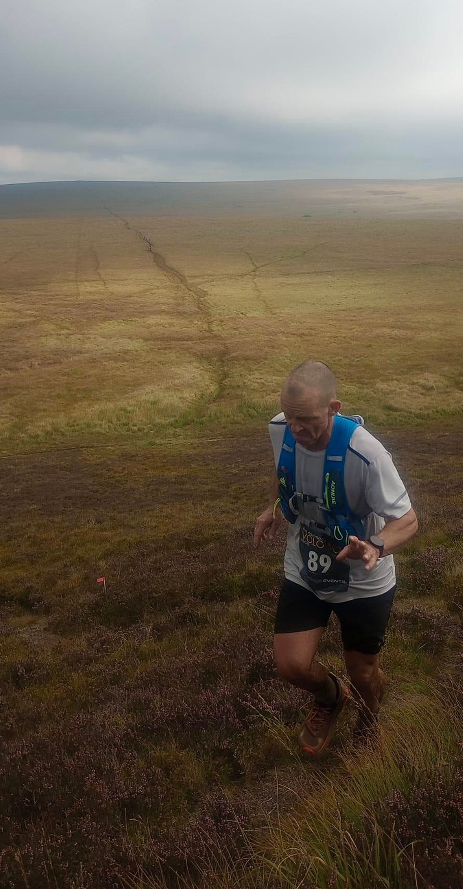 A person wearing a race bib number 89, a hydration vest, and running gear is seen running uphill on a grassy terrain. The background features a vast expanse of open grassy plains under a cloudy sky. The terrain shows a distinct path cutting through the grass.