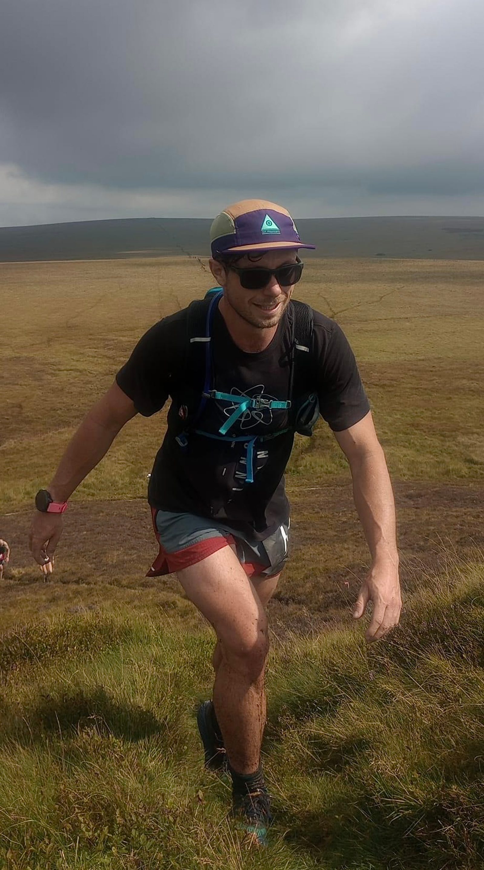 A person with a cap, sunglasses, and a backpack is running uphill on a grassy terrain. The sky is cloudy, and there is a vast open field in the background. The person appears to be participating in an outdoor adventure or trail-running activity.
