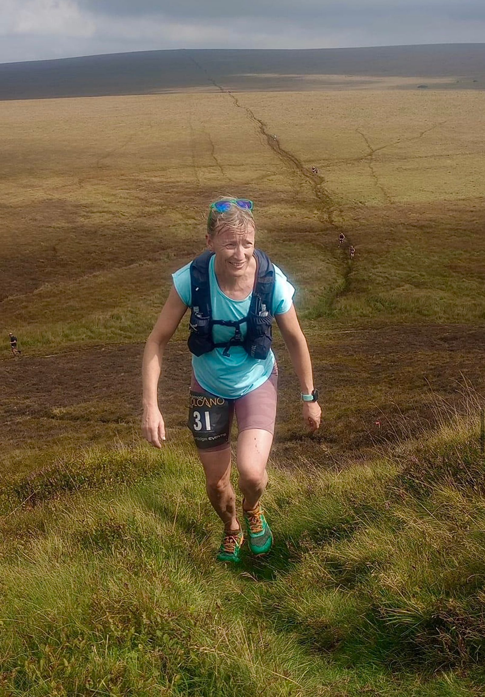 A person wearing a light blue shirt, shorts, and a black running vest with the number 31 is shown ascending a grassy hill. Other runners can be seen in the background on a trail stretching across an expansive field under a cloudy sky.