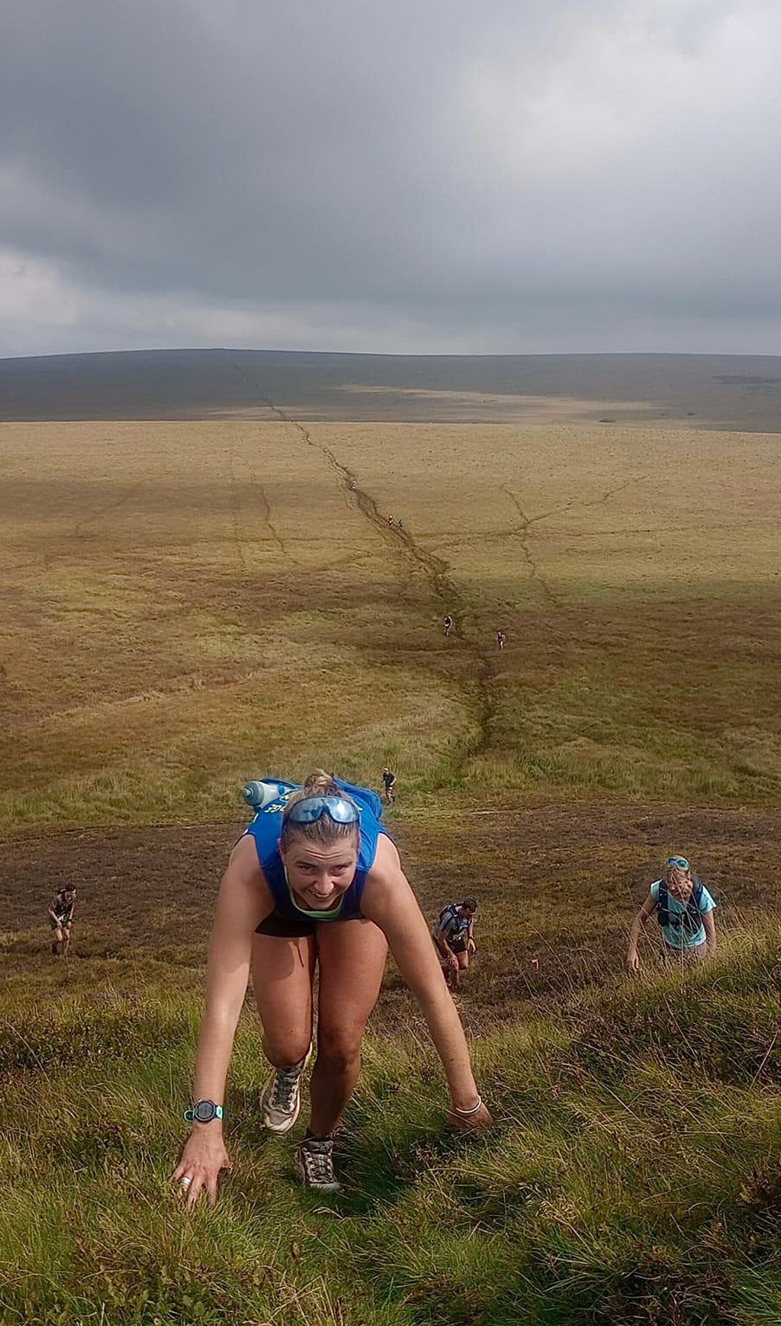 A group of people are hiking up a steep grassy hill. The nearest hiker, wearing a blue tank top and a blue backpack, is crawling up on all fours. Other hikers can be seen in the distance, with vast grassy plains and a cloudy sky in the background.