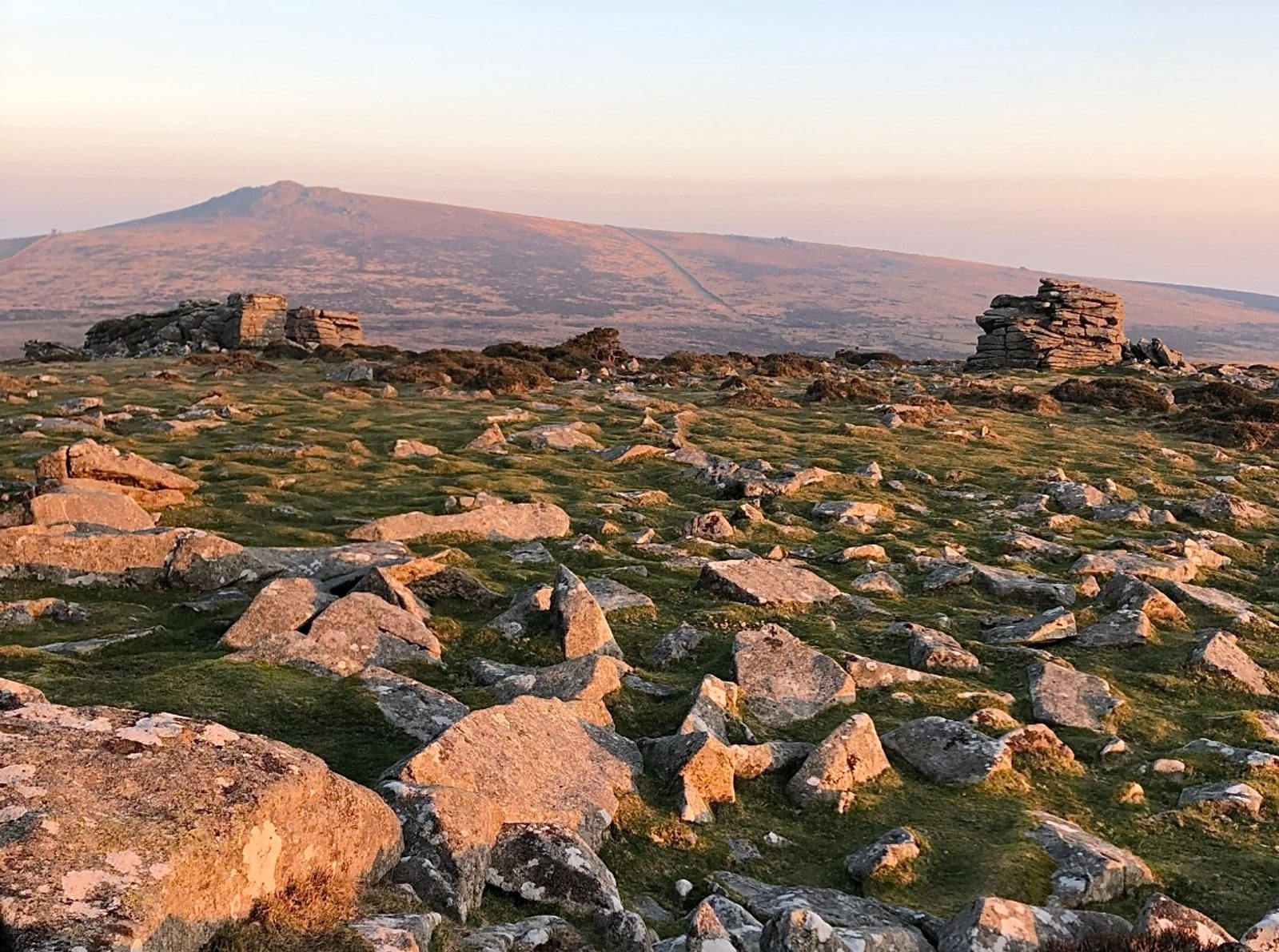 Rocky terrain with scattered green patches under a gentle sunset. In the background, there is a distant mountain with a softly sloping peak. The landscape is calm, and the sky is clear with warm golden hues. Stone structures, likely ancient, are scattered around.
