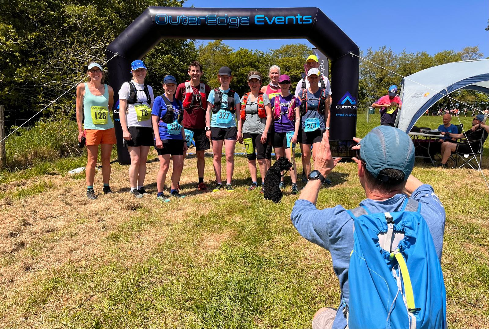 A group of runners pose in front of an "Outeredge Events" inflatable arch. They wear various race numbers and outdoor gear. A person in a blue shirt and cap photographs them from a crouched position, while a small black dog sits nearby.