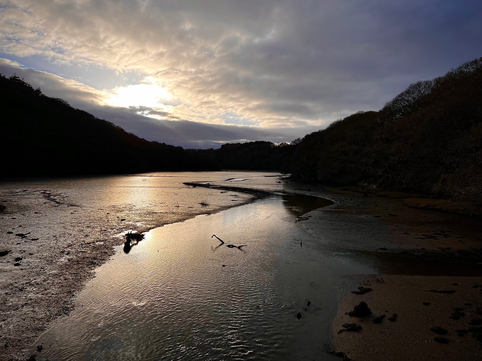 A serene river meanders through a valley at dusk, reflecting the golden hues of the setting sun. Tree-covered hills frame the scene, with dark clouds partially obscuring the sky and leaving patches of warm light on the water's surface.