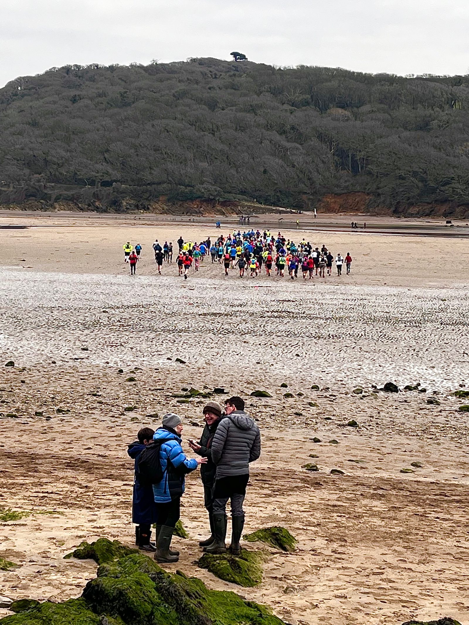 A group of people prepares to run on a sandy beach, moving inland from near the water's edge. In the foreground, a few others stand on the shoreline, some in conversation, with a forested hill in the background under a cloudy sky.