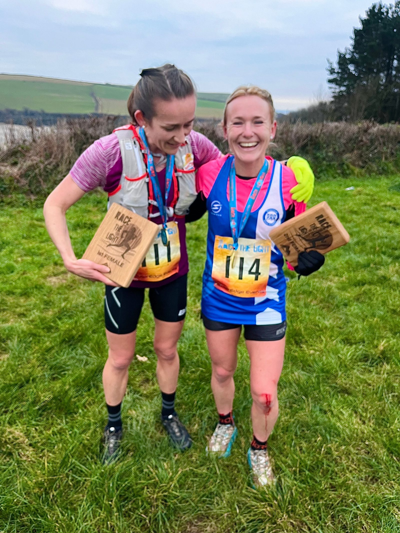 Two women, dressed in athletic attire with race bibs, stand on a grassy field smiling and holding wooden plaques after completing a race. Both wear medals around their necks, and one has a small bandage on her knee. A hilly landscape and cloudy sky are in the background.