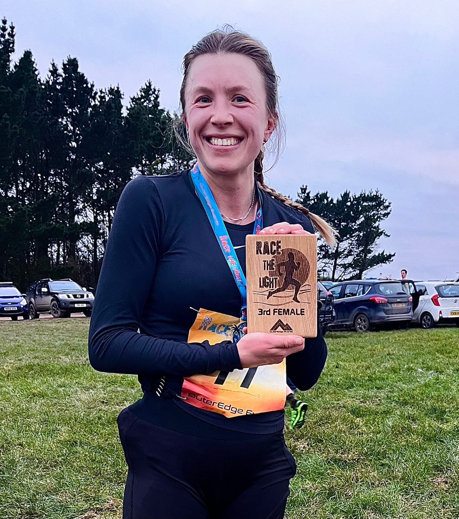 A woman stands outdoors, smiling and holding a wooden plaque that says "Race the Light 3rd Female." She is wearing a black top, a race bib, and a medal around her neck. Cars are parked in the background, and trees can be seen behind them.