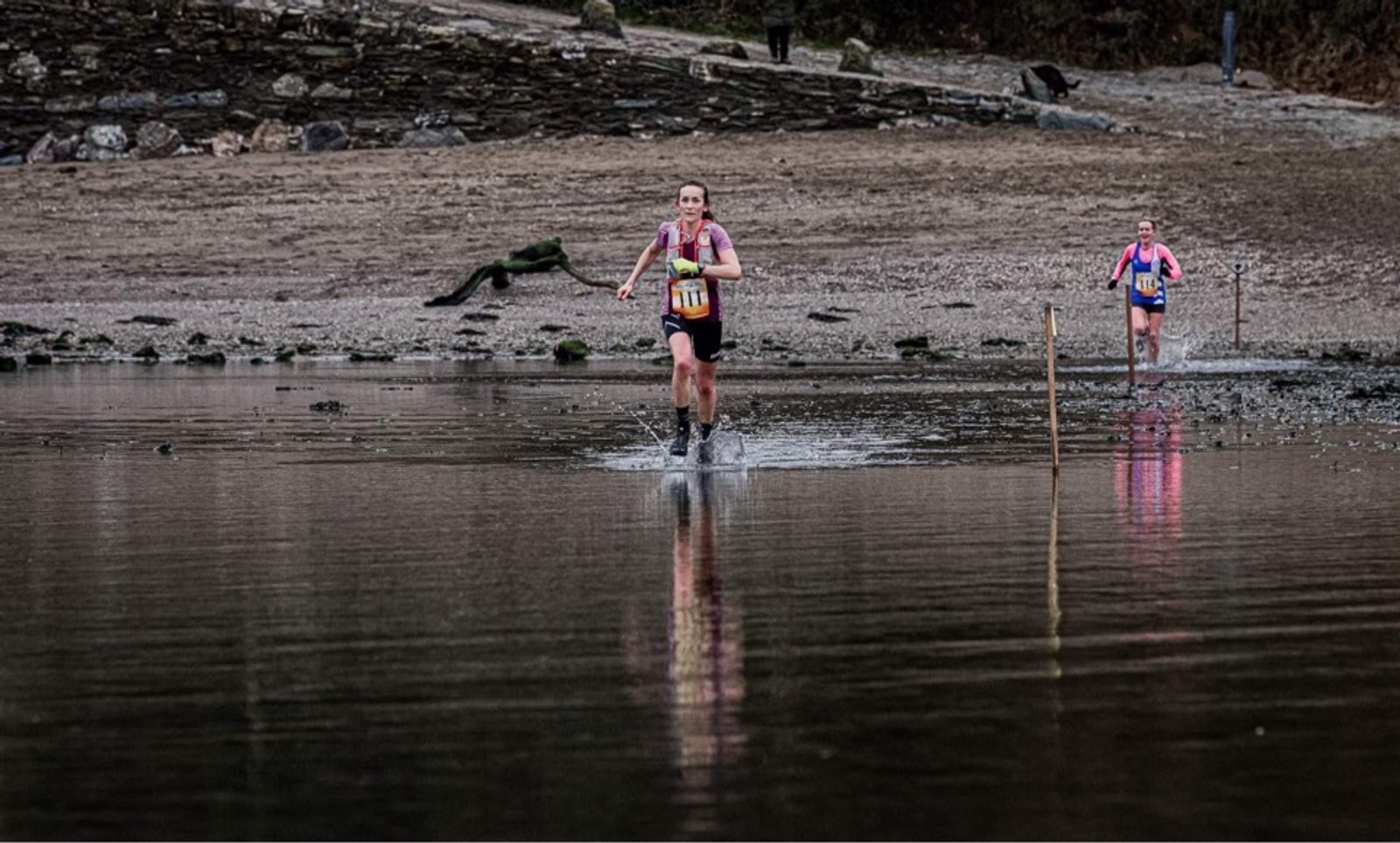 Two athletes in race bibs run through shallow water on a beach, splashing as they go. The first runner is slightly ahead, and the second follows closely behind. The beach is rocky and slopes upwards away from the water. The sky is overcast.