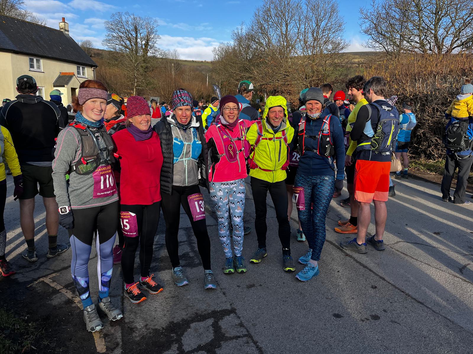 A group of six runners in colorful athletic gear pose for a photo before a race. They stand on a road with other participants and spectators in the background. Leafless trees and a cottage are visible under a sunny sky. .