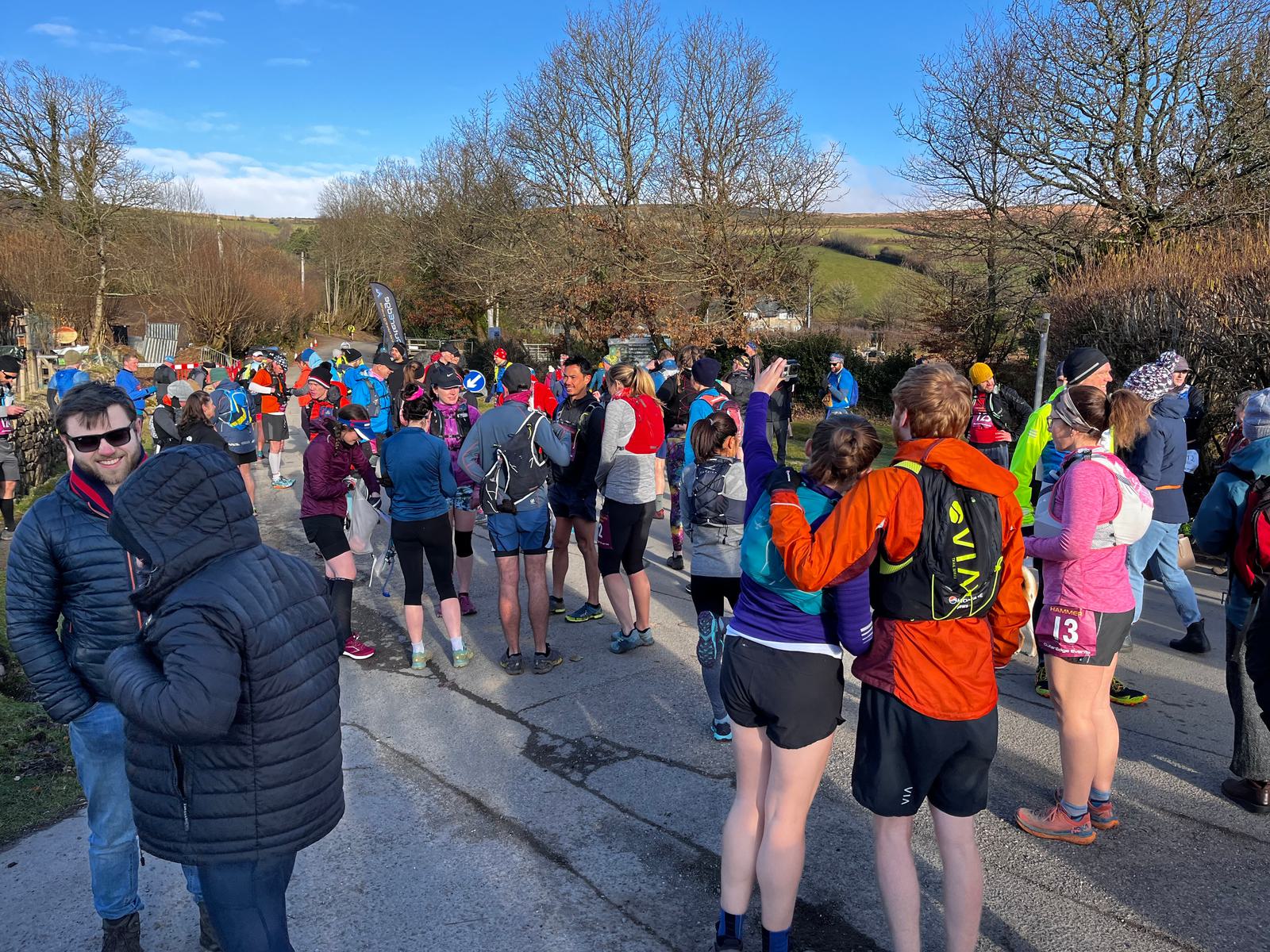 A group of people, many wearing athletic gear and backpacks, gather outdoors on a sunny day. Some are stretching, while others chat in small groups. Trees and rolling hills are visible in the background, suggesting a rural setting.