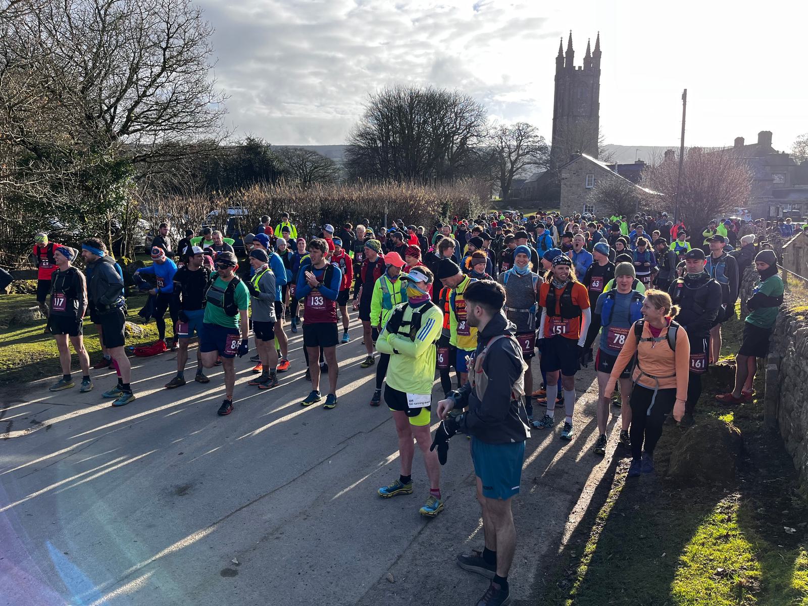 A large group of runners gathers at the starting line of an outdoor race on a clear day. Participants are dressed in colorful athletic gear and are numbered. A prominent church tower and trees are visible in the background against a partly cloudy sky.