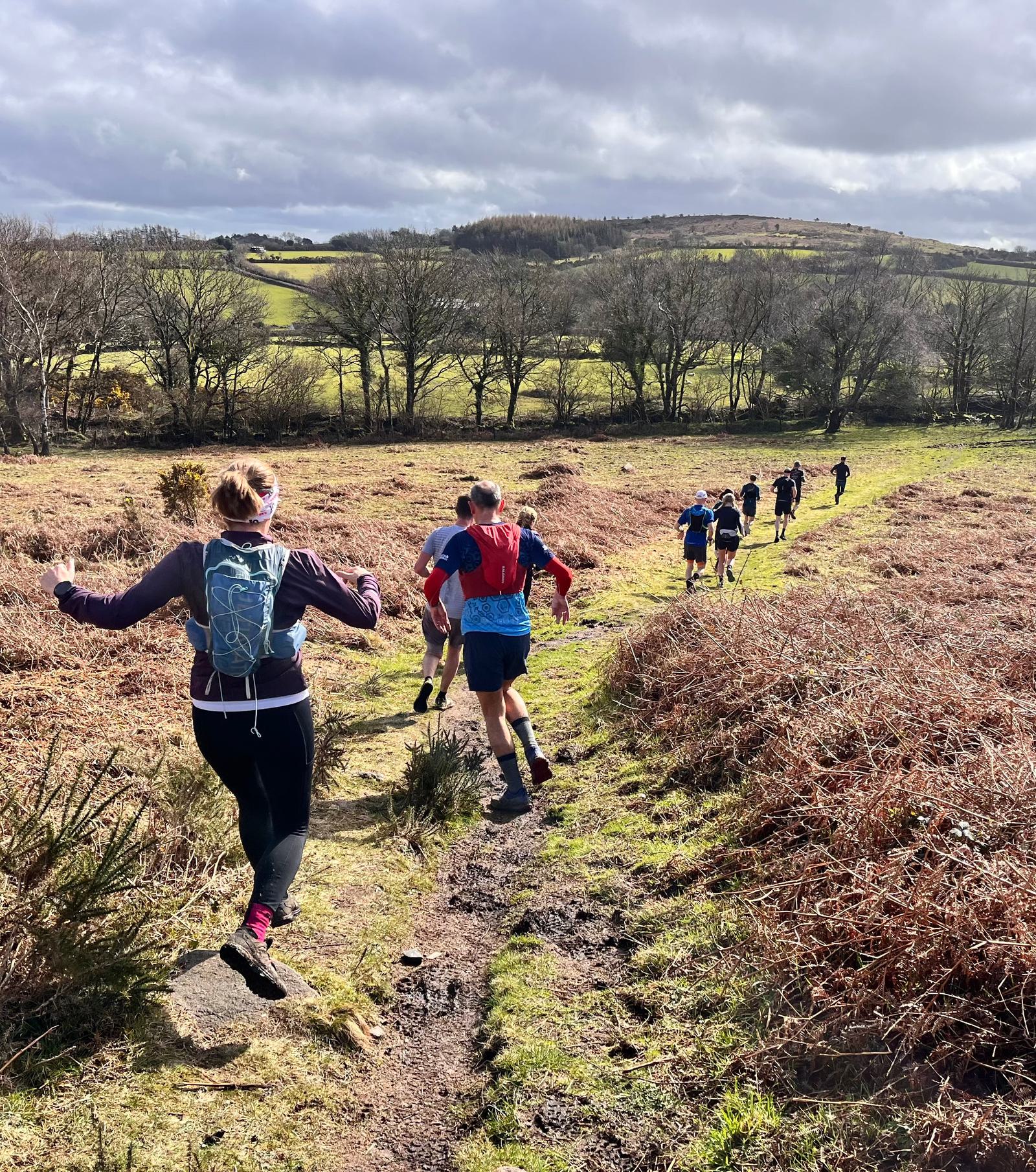 A group of people trail running on a narrow path through a scenic, hilly landscape. They are wearing casual athletic clothing, backpacks, and the area is surrounded by dry grass and trees under a cloudy sky.