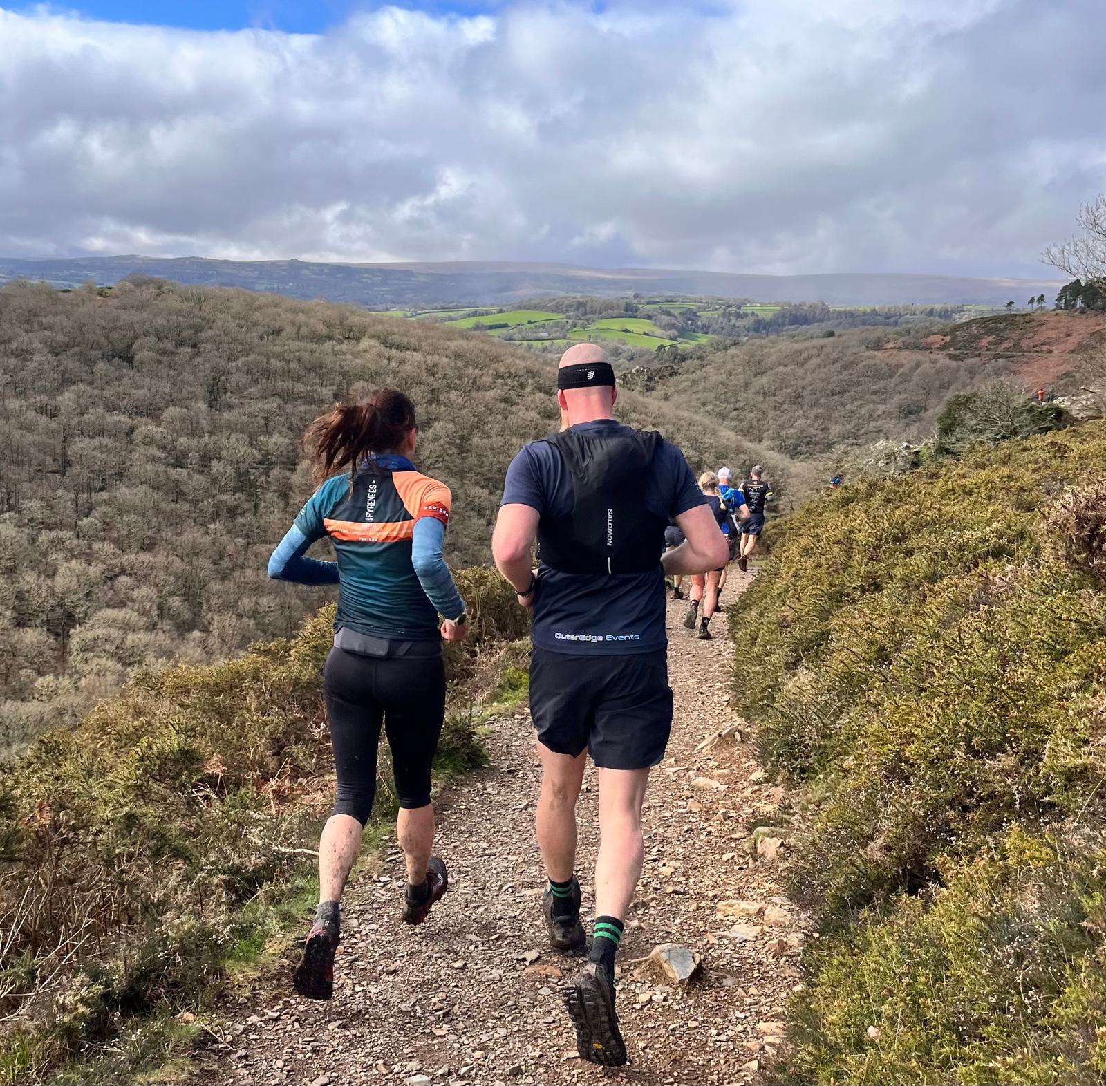 Two people in athletic gear run on a narrow, rocky trail through a hilly landscape. Green hills and cloudy skies stretch into the distance, while other runners are visible further along the path.