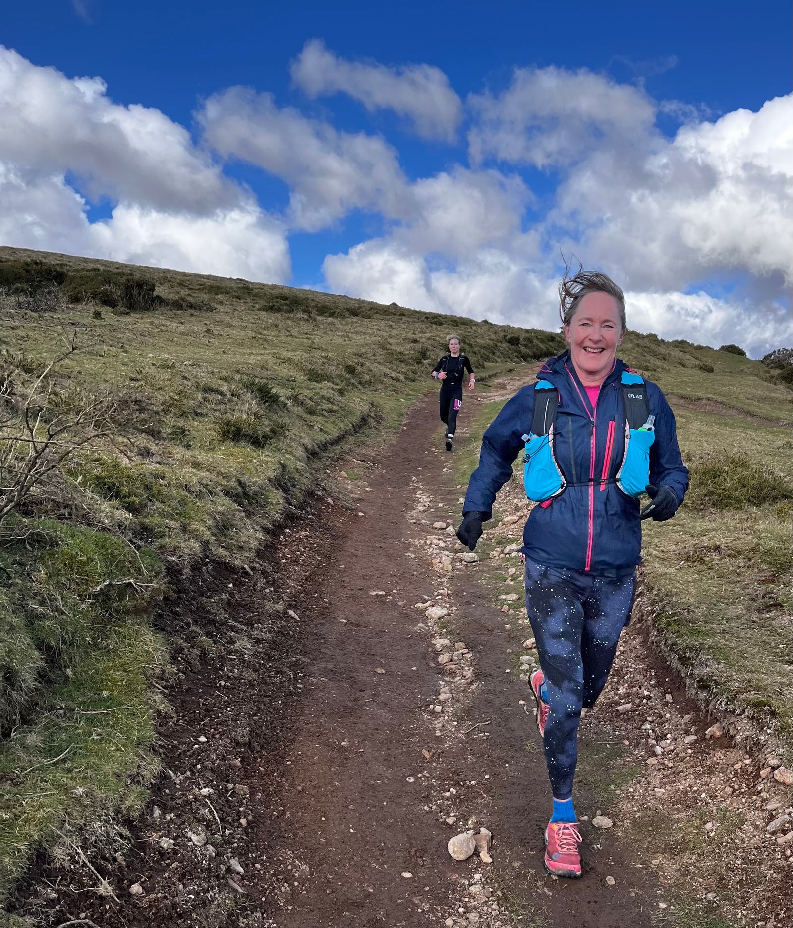Two people are trail running on a rocky path through a grassy hillside under a partly cloudy sky. The person in the foreground is smiling, wearing a blue jacket, a turquoise hydration pack, and pink running shoes, while the person in the background follows behind.