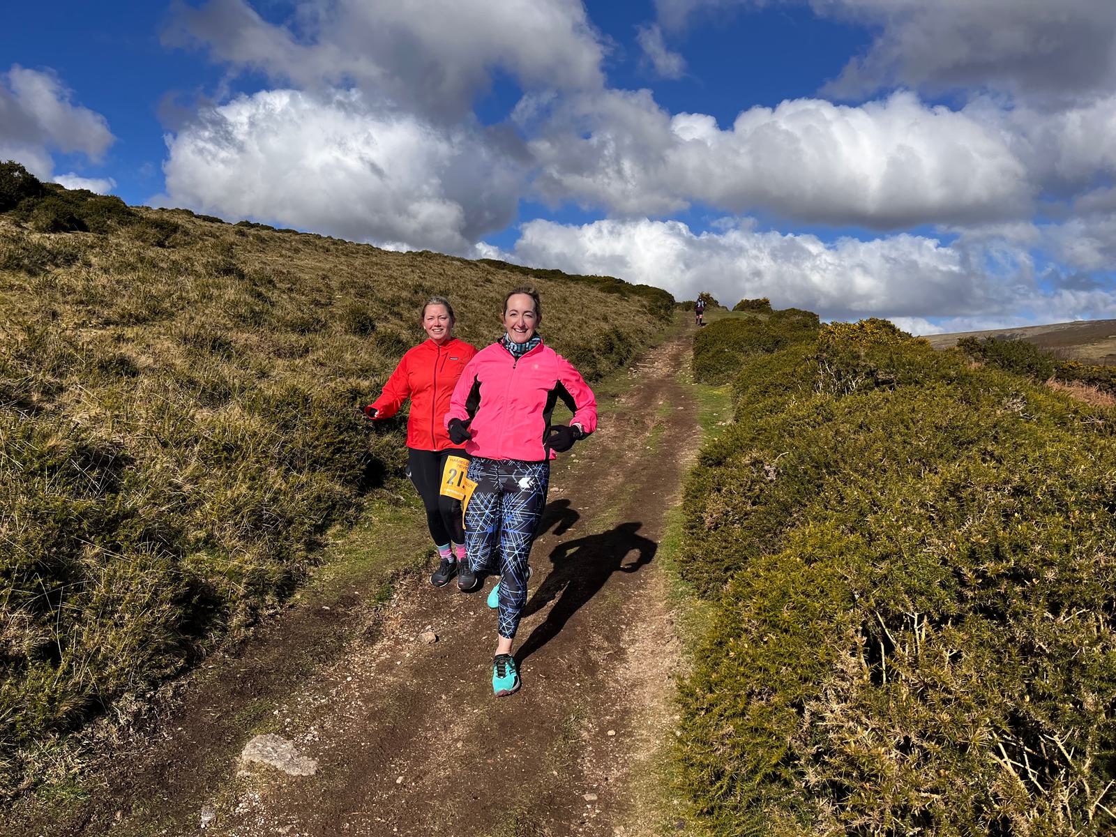 Two women are running on a scenic trail surrounded by greenery under a partly cloudy sky. They are both wearing vibrant jackets, one in red and the other in pink, and appear joyful and energetic as they run uphill.