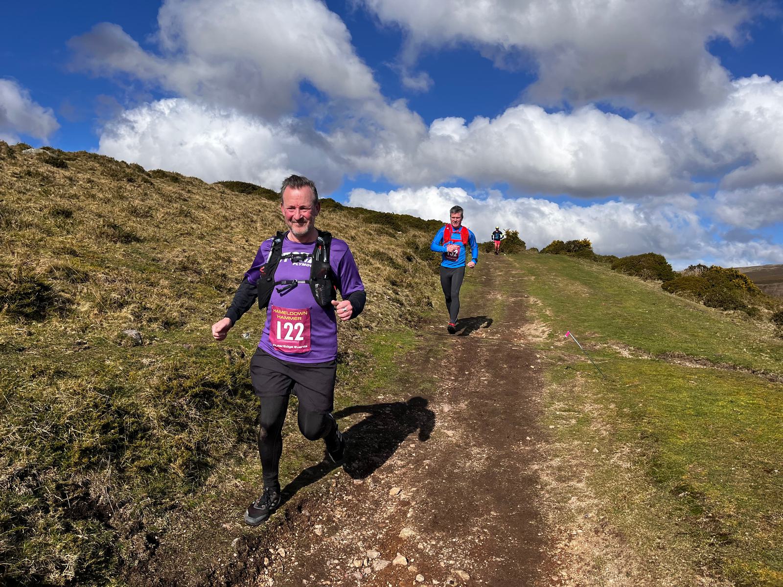 Two men running on a trail during a race on a grassy hillside under a partly cloudy sky. The runner in the foreground wears a purple shirt, black leggings, and a bib with the number 122. The second runner, wearing a blue shirt, is farther back on the path.