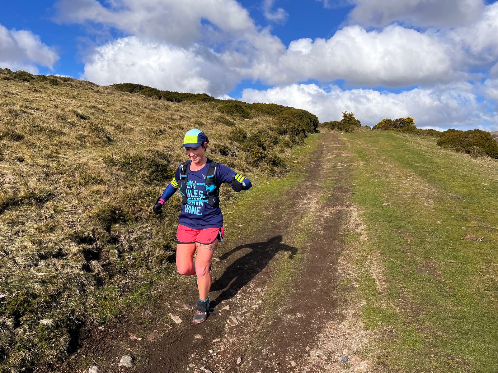 Person wearing a colorful outfit with a cap and a hydration vest running on a dirt path through a grassy landscape. The sky is blue with scattered clouds, and the runner appears to be enjoying the outdoors.