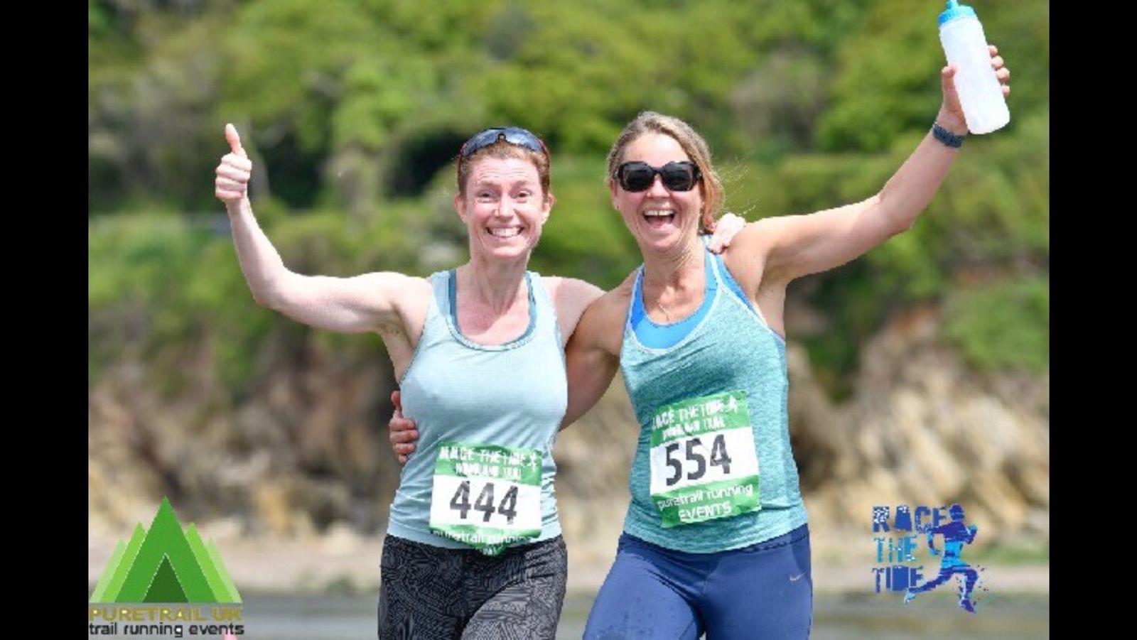 Two women in athletic gear and numbered bibs, smiling and posing with arms around each other after a run. One holds a water bottle. They are outdoors with greenery and rocks in the background. Logos for Puretrail events and "Race the Tide" are visible.