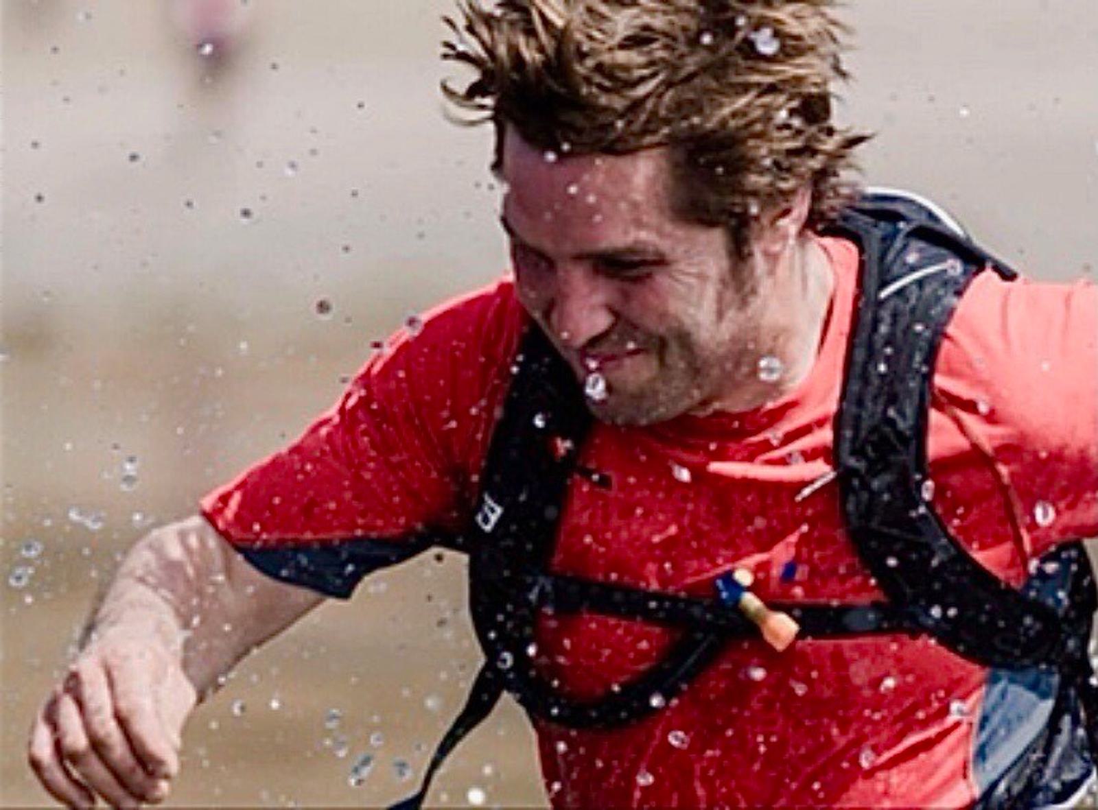 A person with wet hair, wearing a red shirt and a backpack, runs through splashing water. The background is blurry, suggesting movement and speed.