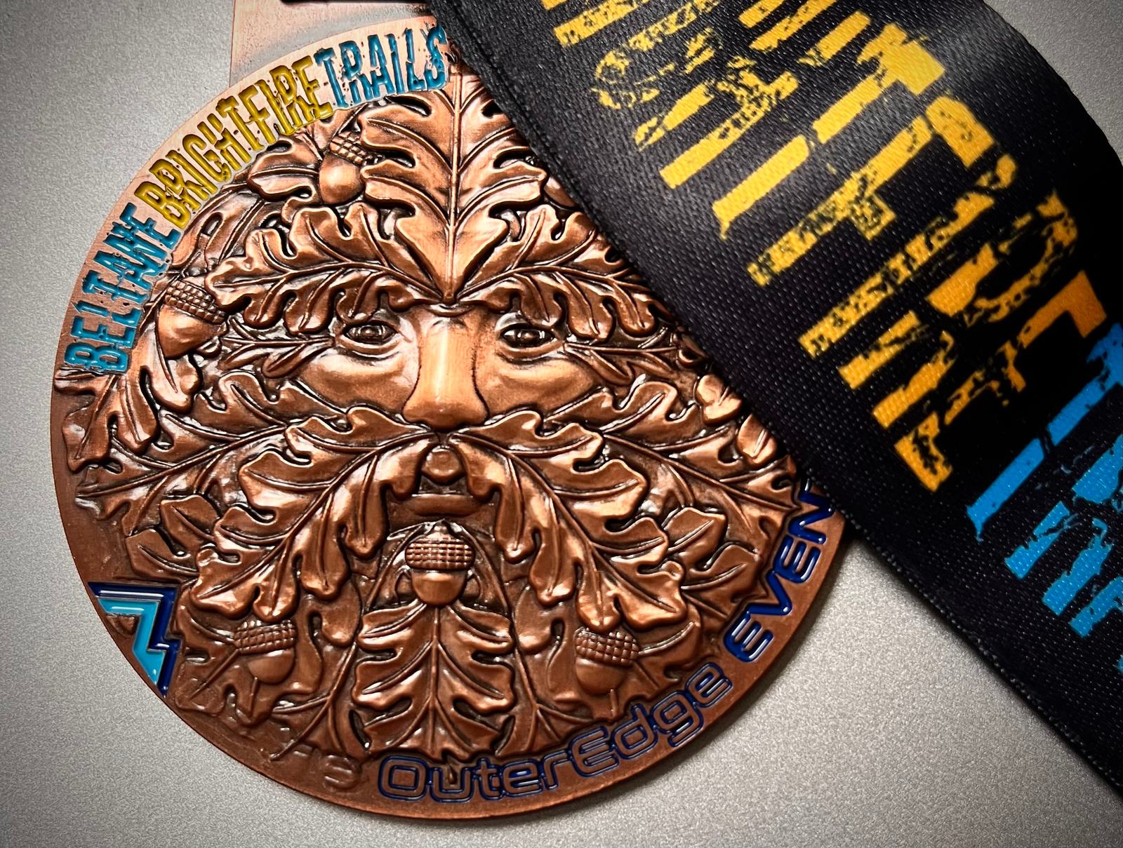 A bronze medal featuring a detailed depiction of a face with leaves and acorns. The text on the medal reads "Beltane Brighter Trails" and "OuterEdge Events." A black ribbon with yellow and blue text is attached.