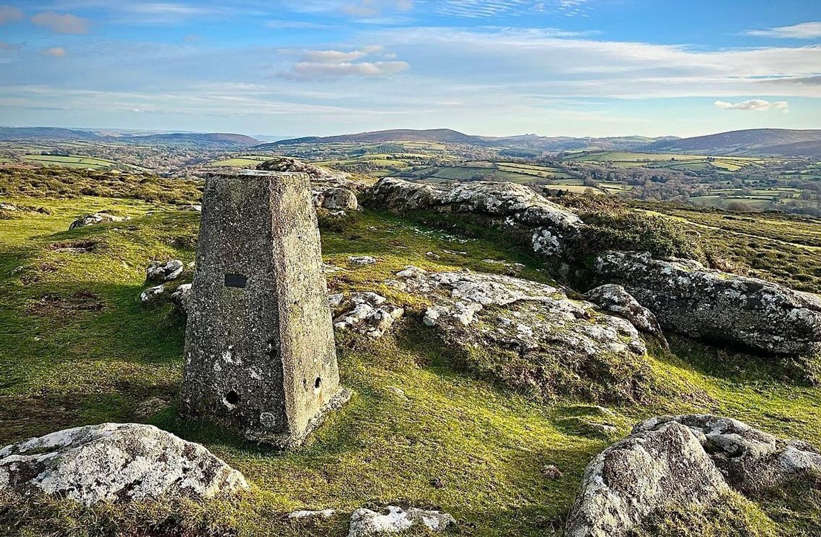 A weathered trig point pillar stands on a grassy, rocky landscape with expansive views of rolling hills and valleys under a sky dotted with clouds. The distant hills are covered in patches of green fields and hedgerows.