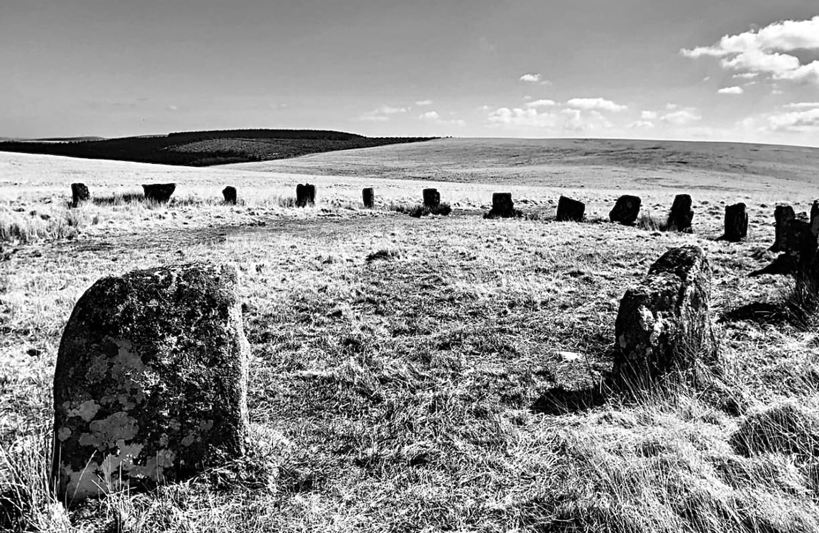 A black and white photo of a stone circle in a grassy landscape. Large, weathered stones form a roughly circular pattern, with smaller stones scattered around. The horizon includes rolling hills with a patchy treeline and a sky with scattered clouds.