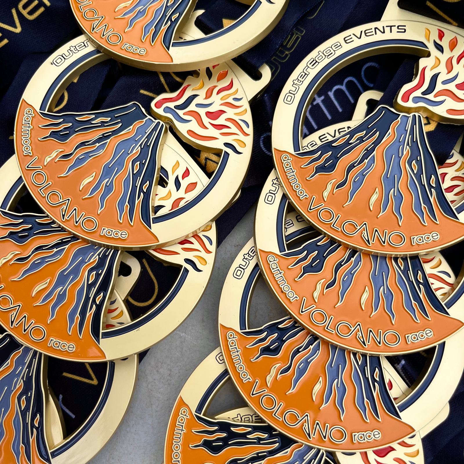 Close-up of a collection of gold and orange medals featuring a volcano design. The text reads "Volcano Race" and "Outrage Events." The medals are stacked on dark blue ribbons with visible fiery patterns.
