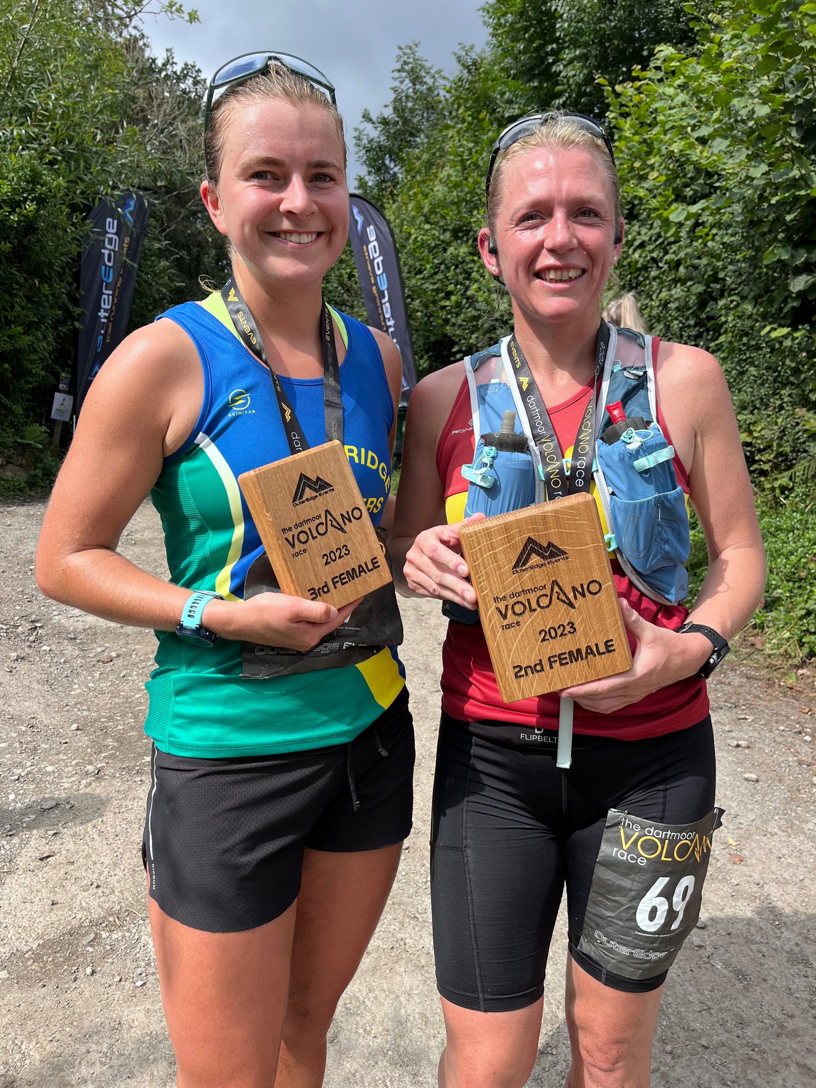 Two women smiling and holding wooden plaques, standing outdoors. Both wear sports attire and medals around their necks. The plaques read "Volcano Race 2023, 2nd Female" and "3rd Female." Trees and race banners are visible in the background.