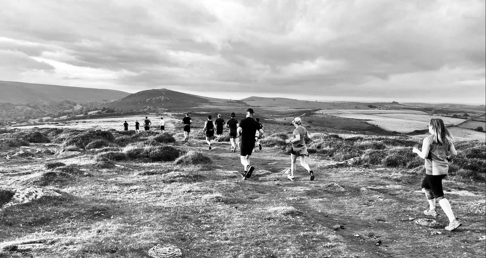 A group of people dressed in athletic wear are seen running on a rocky, uneven path across a hilly, open landscape. The sky is overcast, and the terrain is dotted with patches of grass and shrubs. The scene is in black and white.