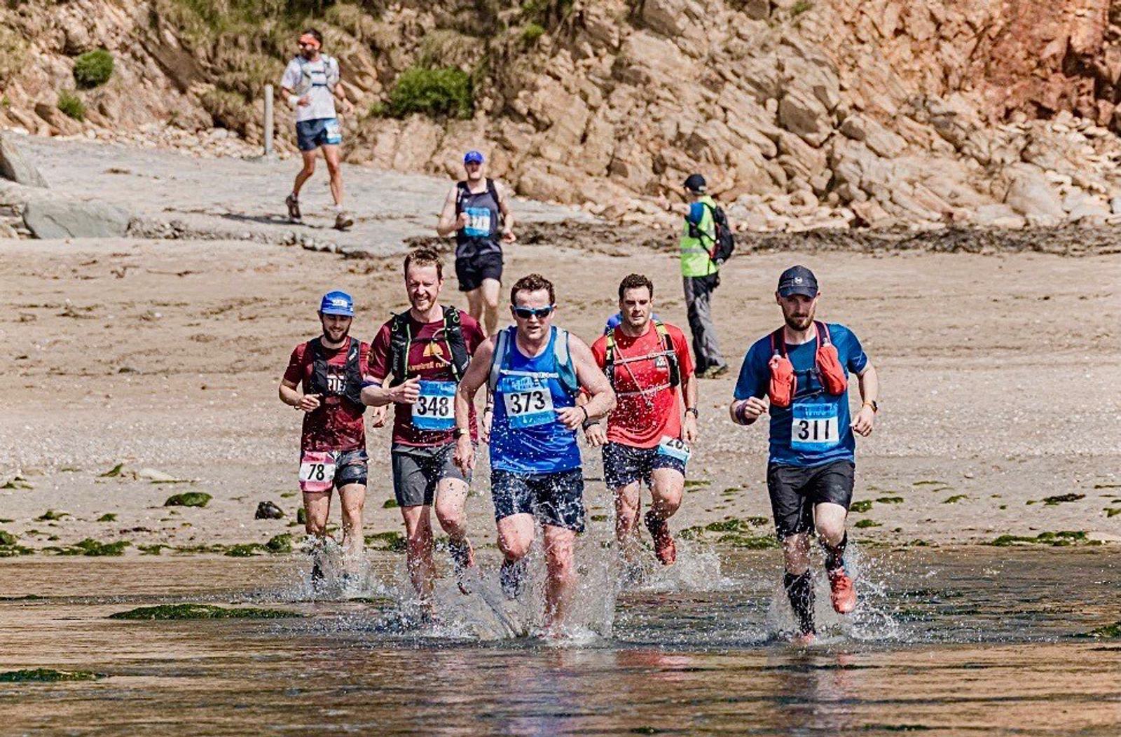 A group of six runners, all wearing race bibs, are running through shallow water on a beach. They are dressed in athletic gear and appear focused. The background features rocky cliffs and sandy terrain.