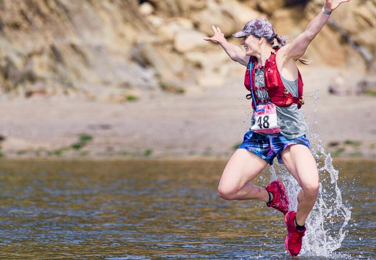 A person in a colorful running outfit and cap joyfully leaps through a shallow body of water. They wear a race bib with the number 48, and splashes rise around them. The background features a rocky shore.