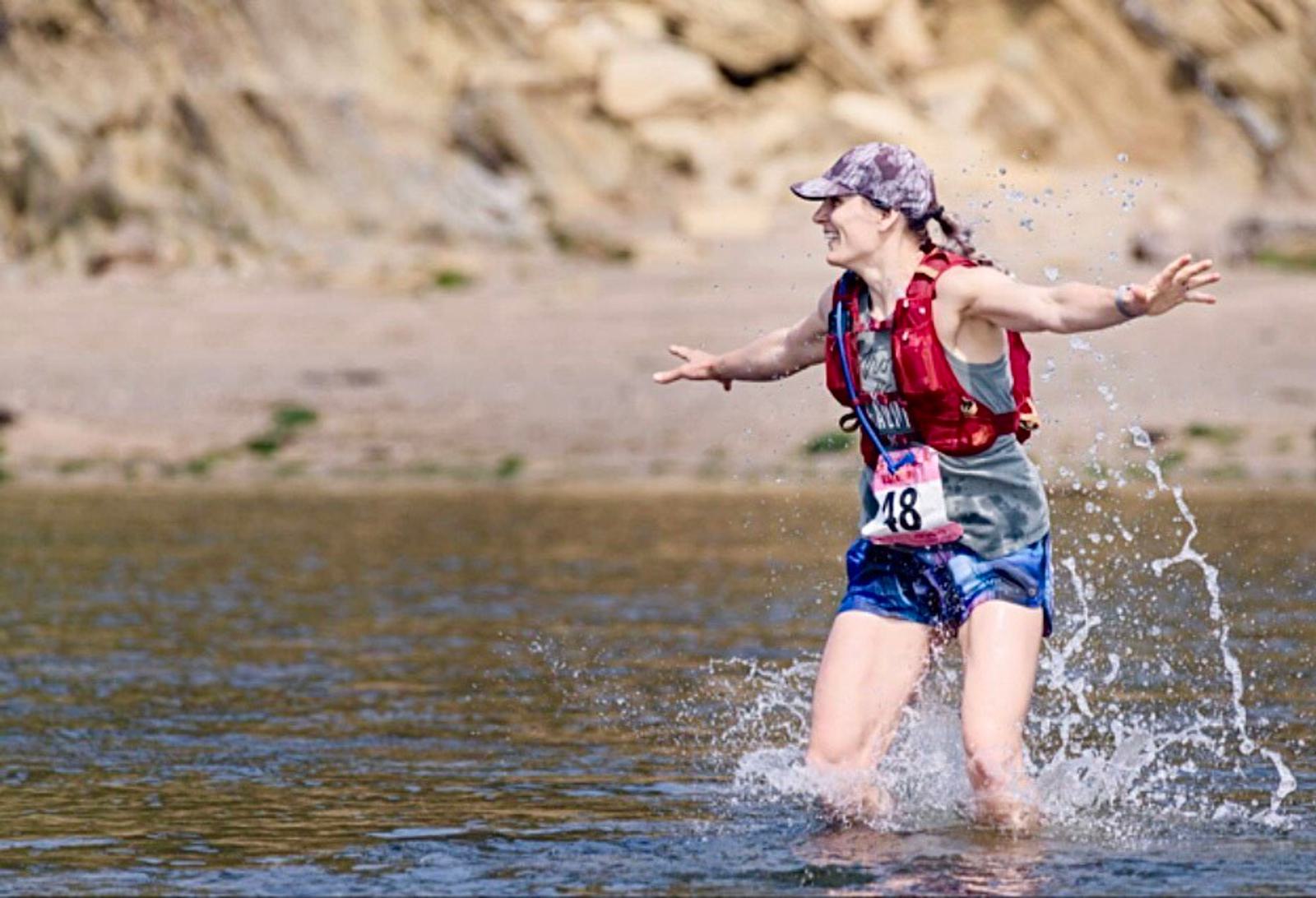 A person wearing a hat, vest, and shorts splashes through a shallow body of water, smiling. They have a race number pinned to their vest. The background features rocky terrain and dry soil.
