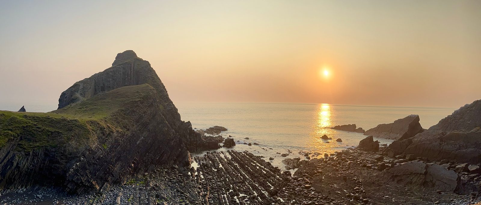 A panoramic view of a rocky coastline at sunset. The sun hangs low in the sky, casting a warm golden glow on the water. The scene features rugged cliffs and a tide pool in the foreground, creating a serene and picturesque landscape.