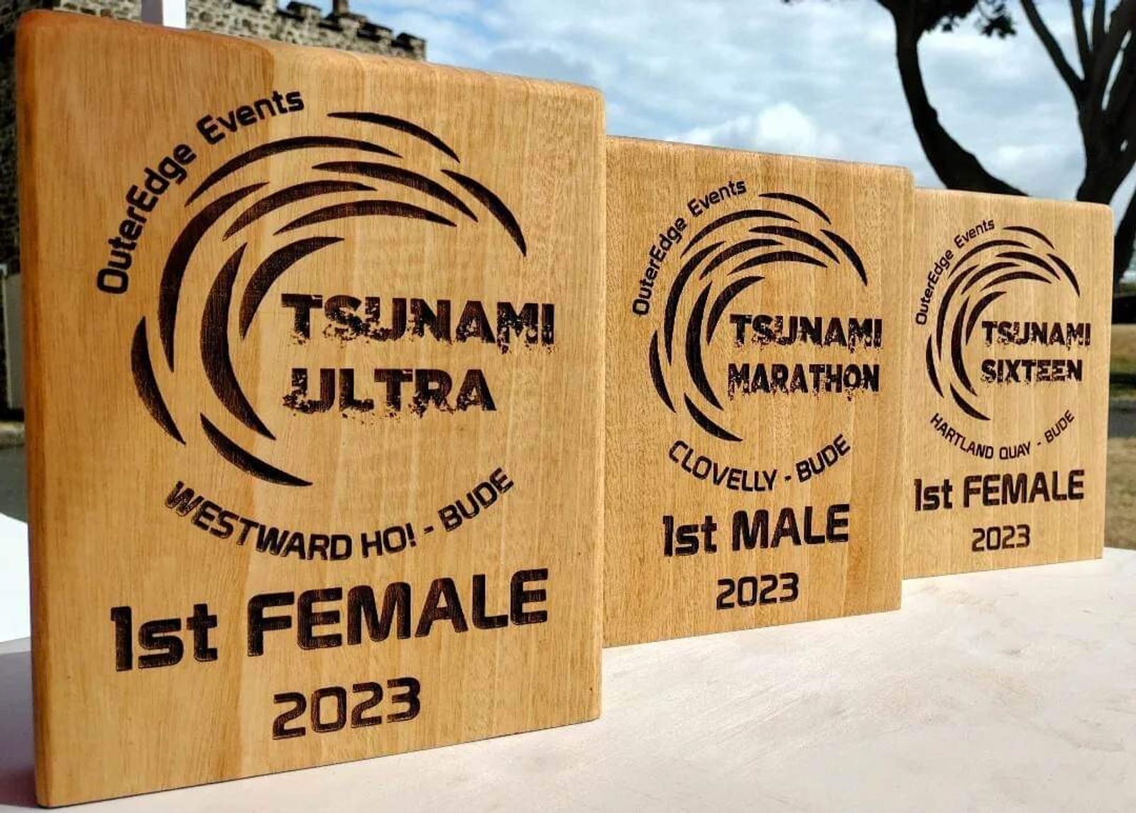 Three wooden plaques showcasing awards from OuterEdge Events are displayed side by side: "Tsunami Ultra 1st Female 2023," "Tsunami Marathon 1st Male 2023," and "Tsunami Sixteen 1st Male 2023." The plaques feature a wave design and the event locations.