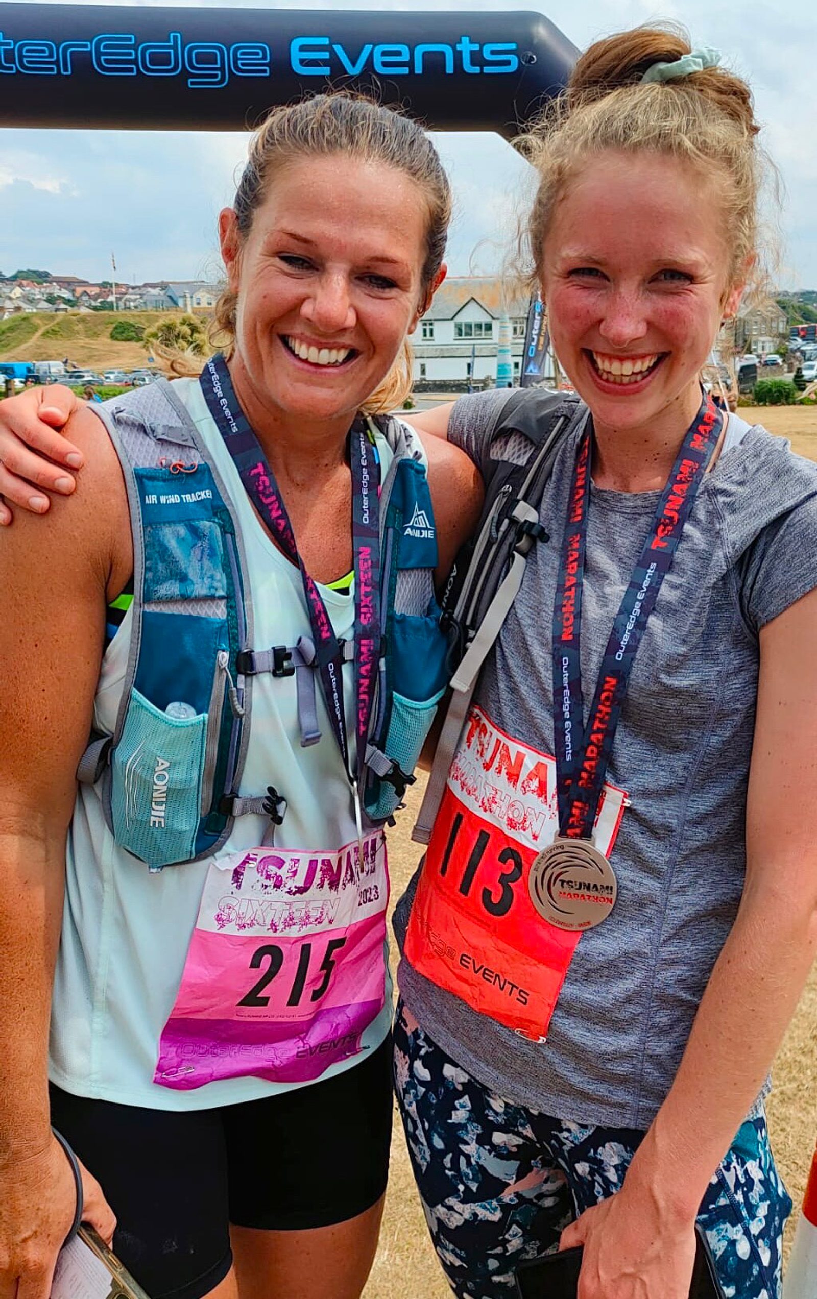 Two smiling women wearing running gear, hydration packs, and event bibs (numbers 215 and 113) pose together at a race finish line. They both have medals around their necks and appear happy and triumphant. An event banner is visible in the background.