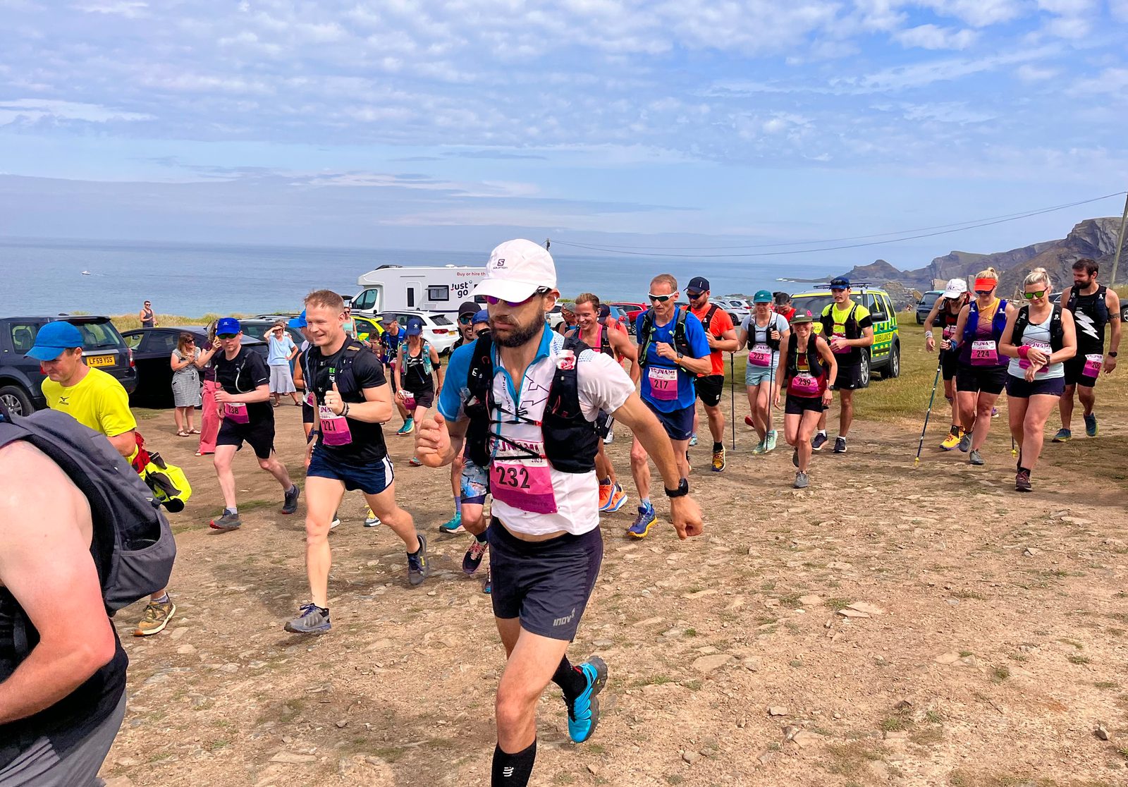 A group of runners participating in an outdoor race along a coastal path. The sky is partly cloudy, and they are wearing running gear with race numbers displayed. A white vehicle and rocky cliffs are visible in the background.