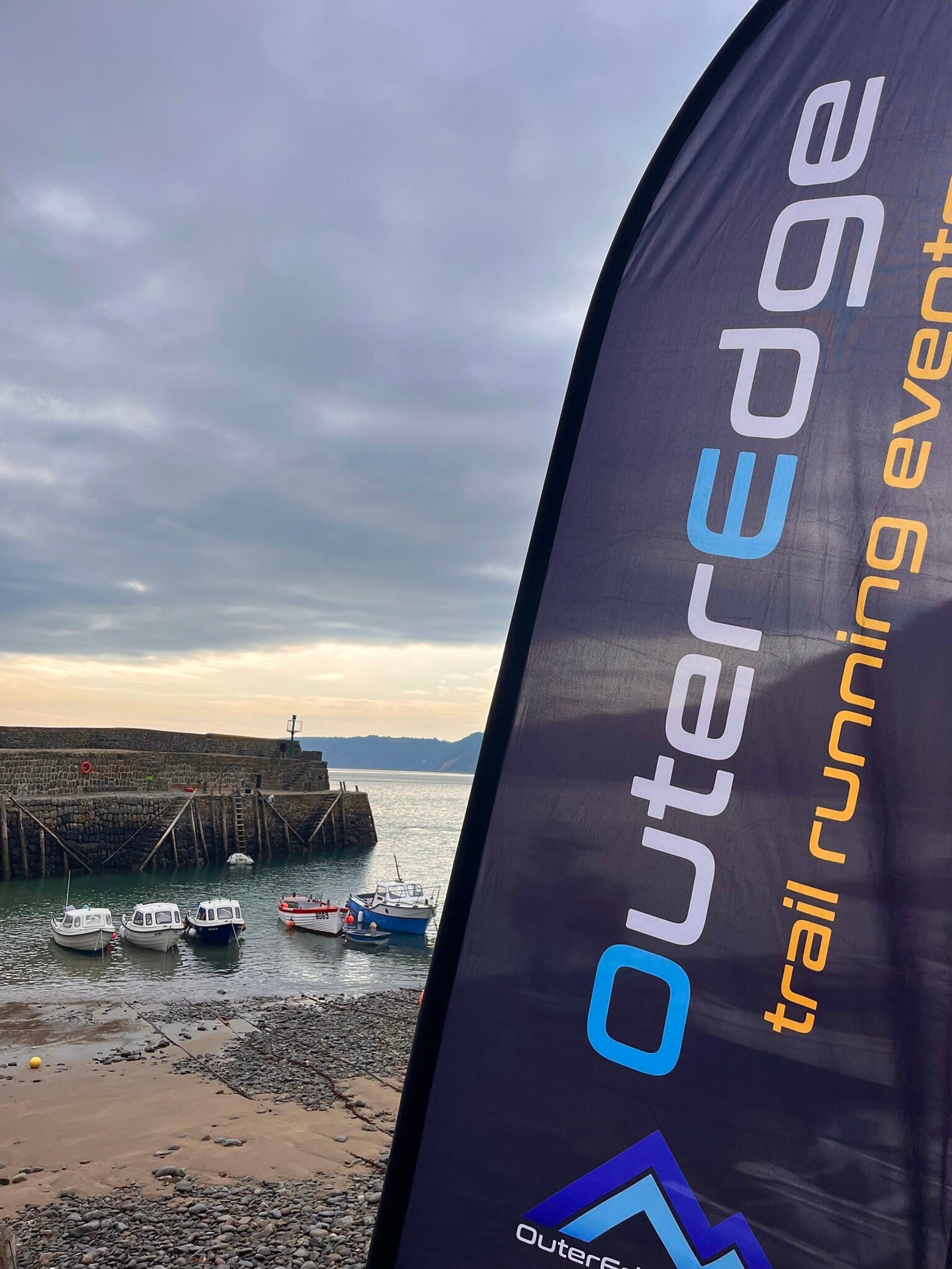 A coastal scene featuring a few small boats docked on a rocky shore, with calm water and cloudy skies in the background. The foreground shows a black flag with the text "OuterEdge trail running events" in white and blue letters.