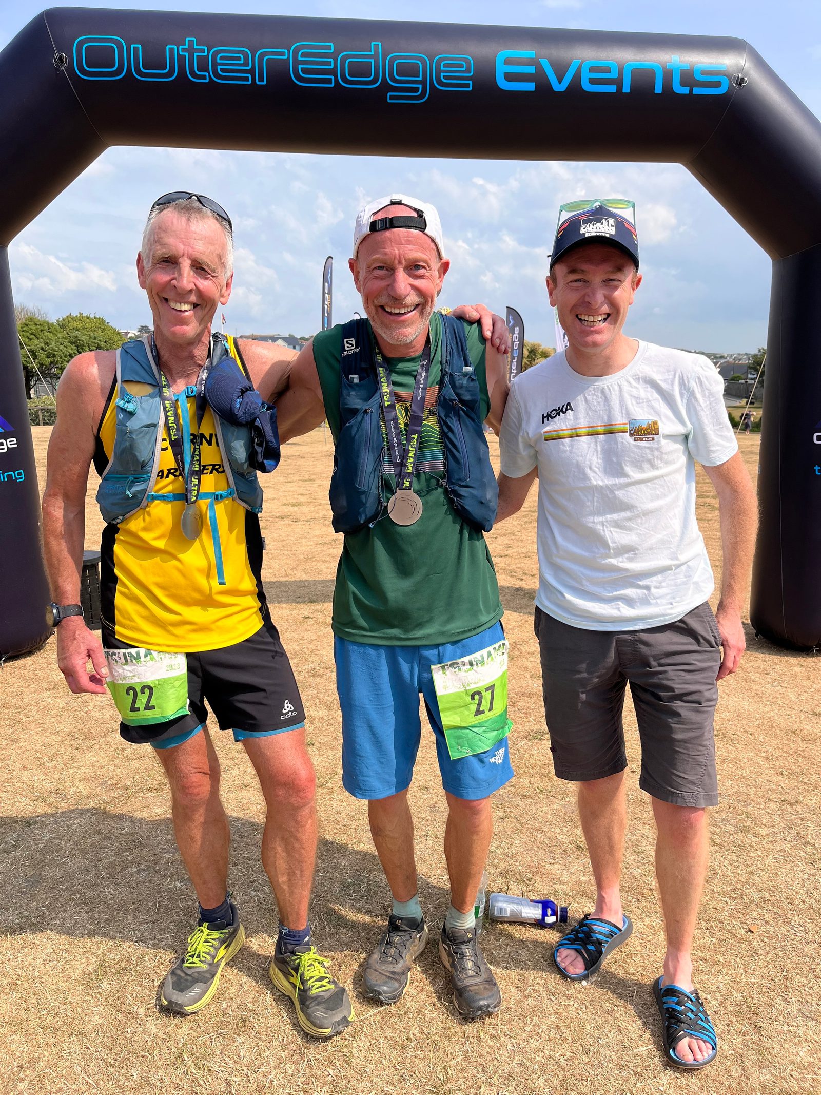 Three men stand smiling under an "OuterEdge Events" banner at a race finish line. Two wear vests and race numbers (22 and 27), one with medals around his neck, while the third is in a white shirt and sandals. The ground is grass, and the sky is partly cloudy.
