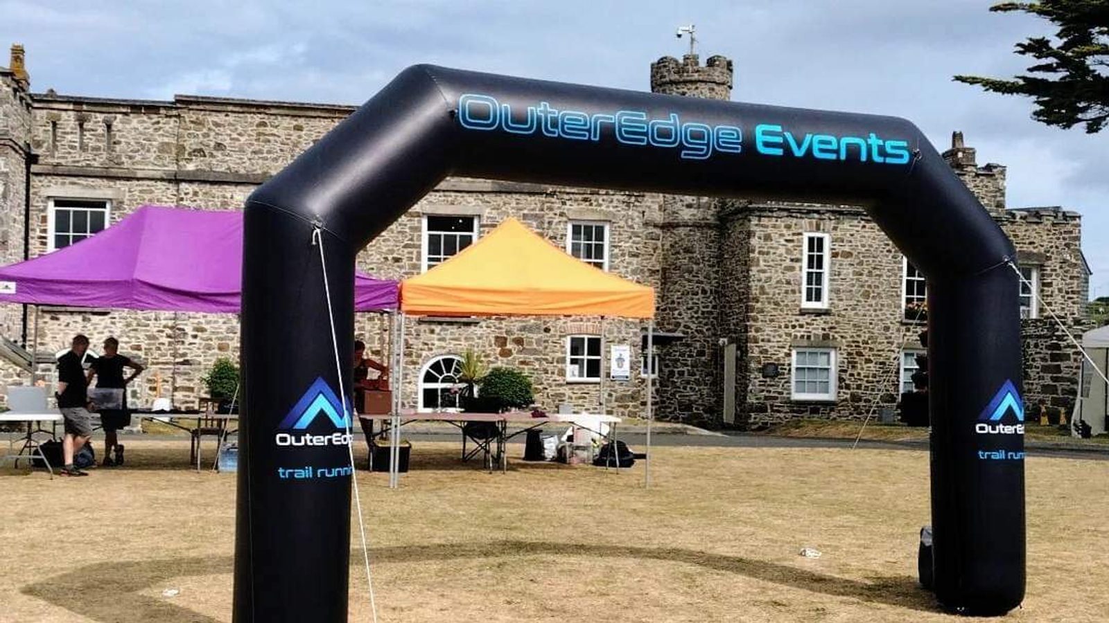 An inflatable arch with the words "OuterEdge Events" marks the entrance to an outdoor event. Beneath it, there are tents with people setting up tables and equipment. The event is set in front of a historic stone building on a partly cloudy day.