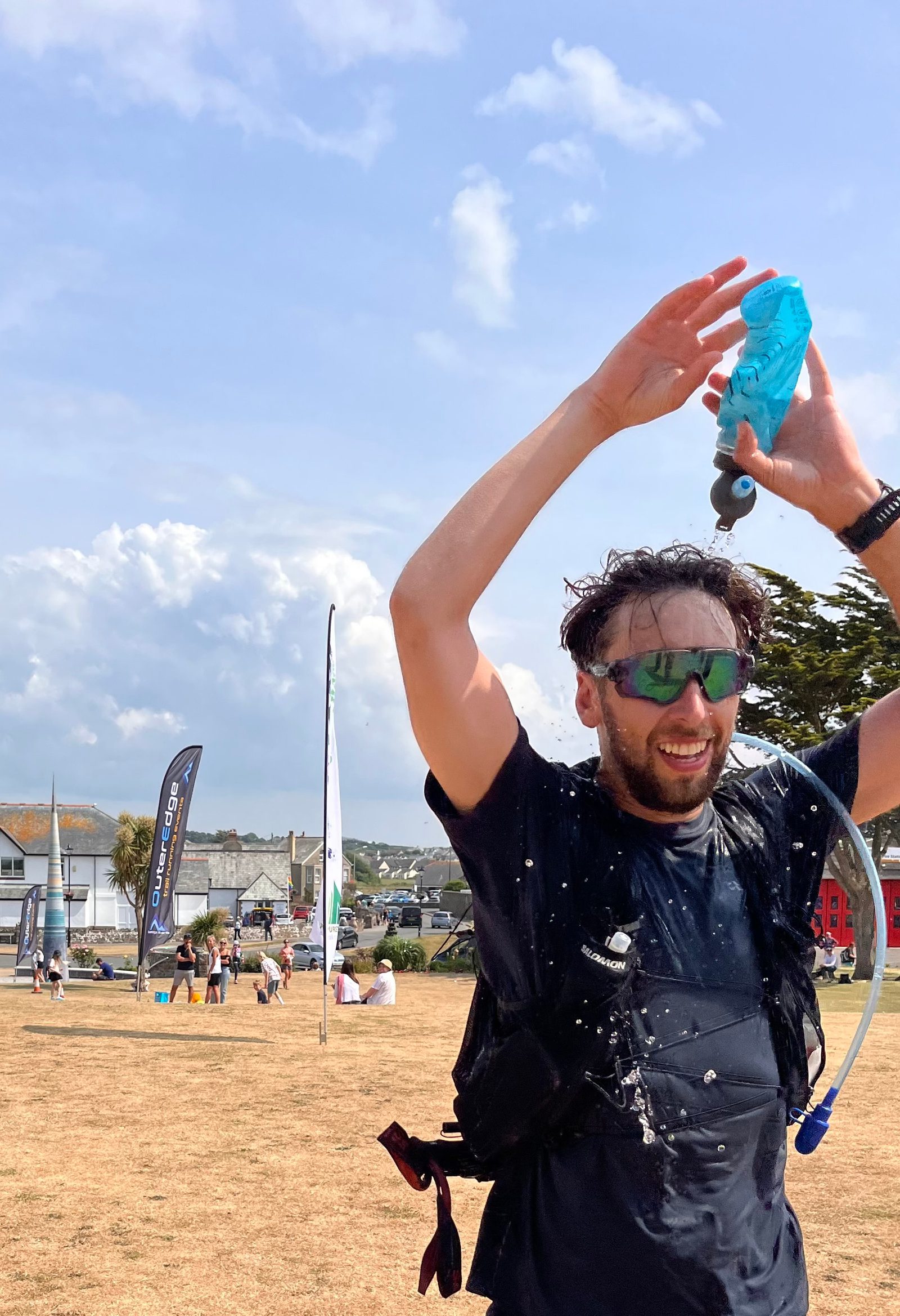 A man in a black t-shirt and sunglasses pours water from a blue bottle over his head on a sunny day. He is wearing a hydration pack with a tube. Behind him is a grassy field with people, flags, and a few buildings in the background.