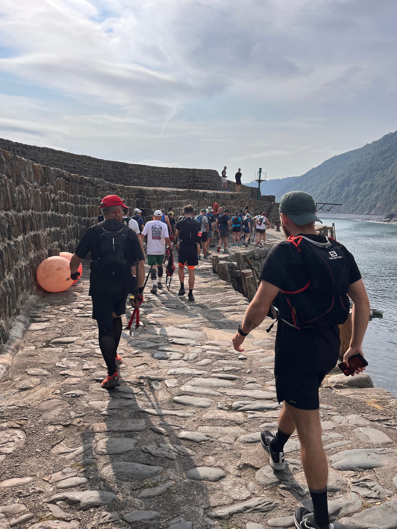 A group of people are running along a cobblestone path beside a body of water, near a stone wall. One person in the foreground carries walking poles and wears a red cap, while others ahead are also wearing athletic gear. Hills and a cloudy sky are in the background.