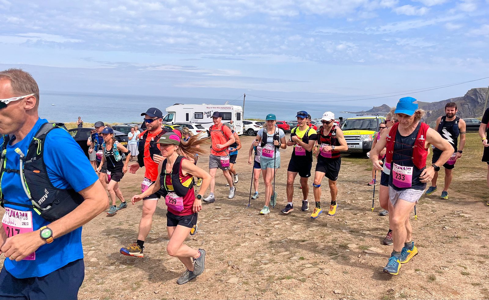 A group of people wearing running gear participate in a trail run near a coastal area. They appear to be in the early stages of the race, running over rocky terrain with the ocean and cliffs in the background. Several vehicles and spectators are visible behind them.