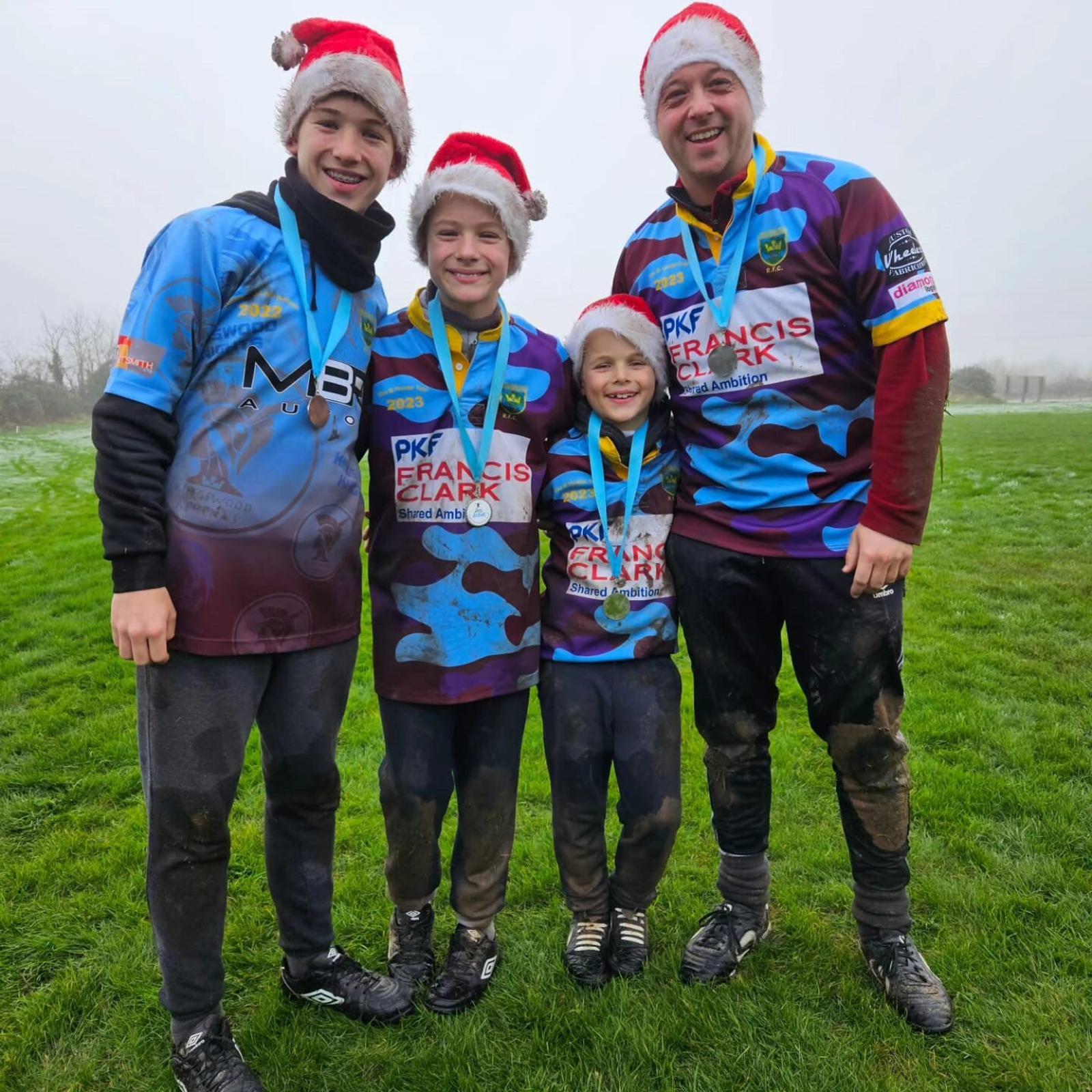 Four people wearing sports jerseys and Santa hats stand on a grassy field, smiling. They are wearing medals around their necks and have mud on their clothes, indicating participation in an outdoor sports event. The background is foggy.