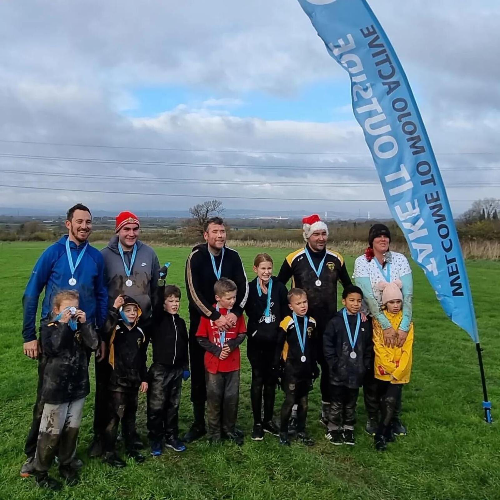 A group of children and adults, some wearing medals, stand on a grassy field under a cloudy sky. Many are covered in mud. A tall banner on the right displays text. The adults wear festive hats, and everyone appears cheerful. Power lines are visible in the background.