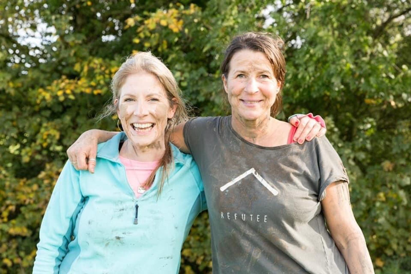 Two women standing outdoors, both smiling and with their arms around each other. They are covered in mud, suggesting they have been participating in an outdoor activity. One wears a light blue hoodie, the other a dark grey shirt with white text. Green trees are in the background.