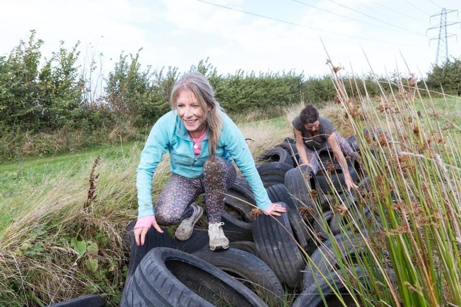 Two women navigate an outdoor obstacle course by climbing over a series of old tires arranged on a grassy field. The woman in the foreground wears a teal jacket and patterned leggings. The scene is set against a backdrop of bushes and a partly cloudy sky.