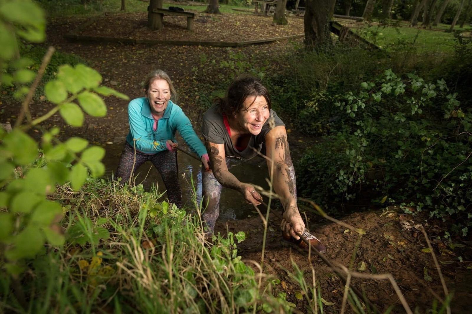 Two women engaged in a muddy, outdoor adventure; one woman, covered in mud, is pulling herself up a short slope with the help of a rope while the other, wearing a light blue top, follows closely behind, both smiling and enjoying the challenge.
.