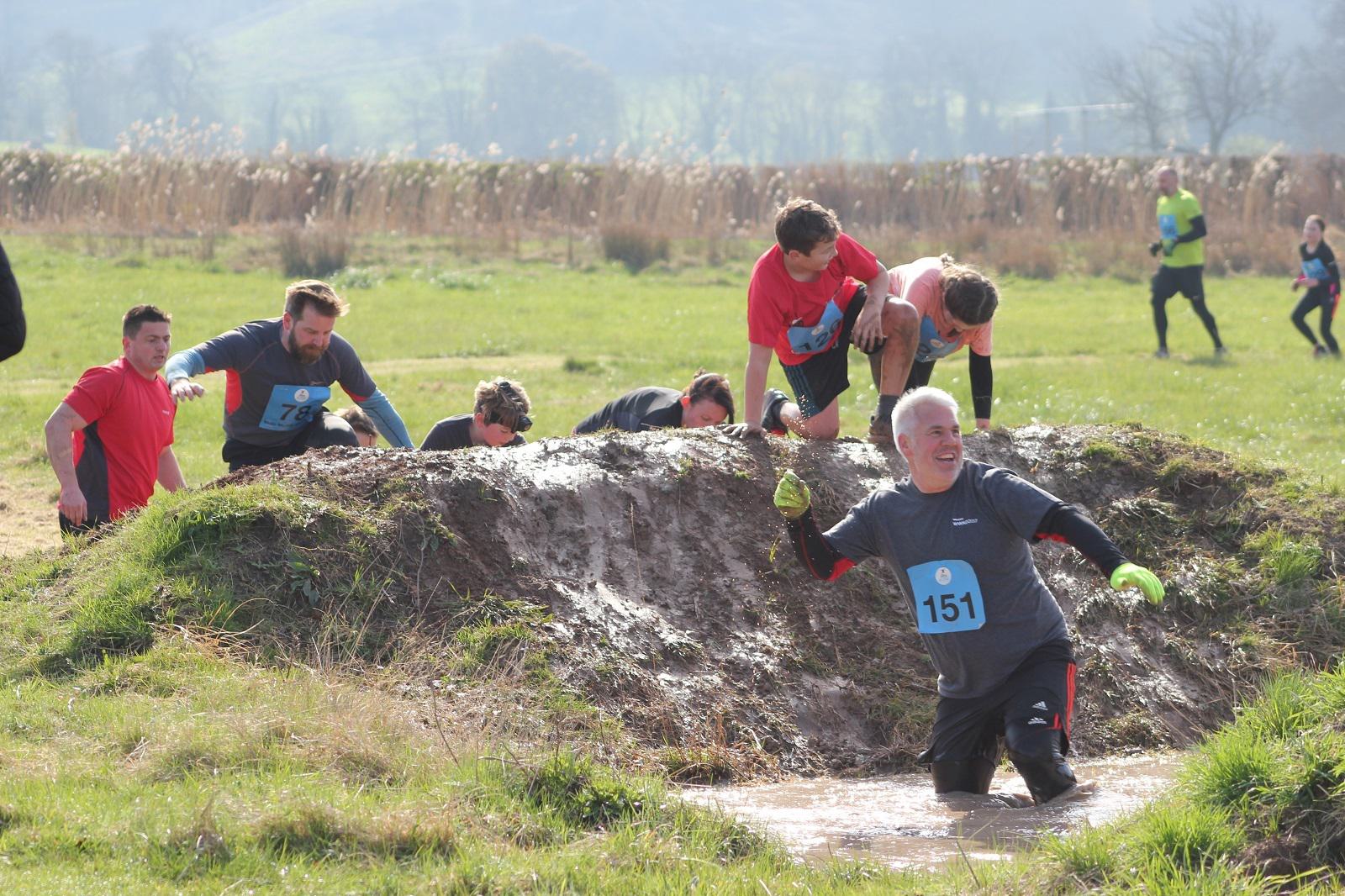 A group of participants in a mud run event navigate a wet and muddy obstacle in an open field. Some are climbing out of a muddy pit while others help their teammates. They wear numbered bibs, and the surrounding area is grassy with hills in the background.