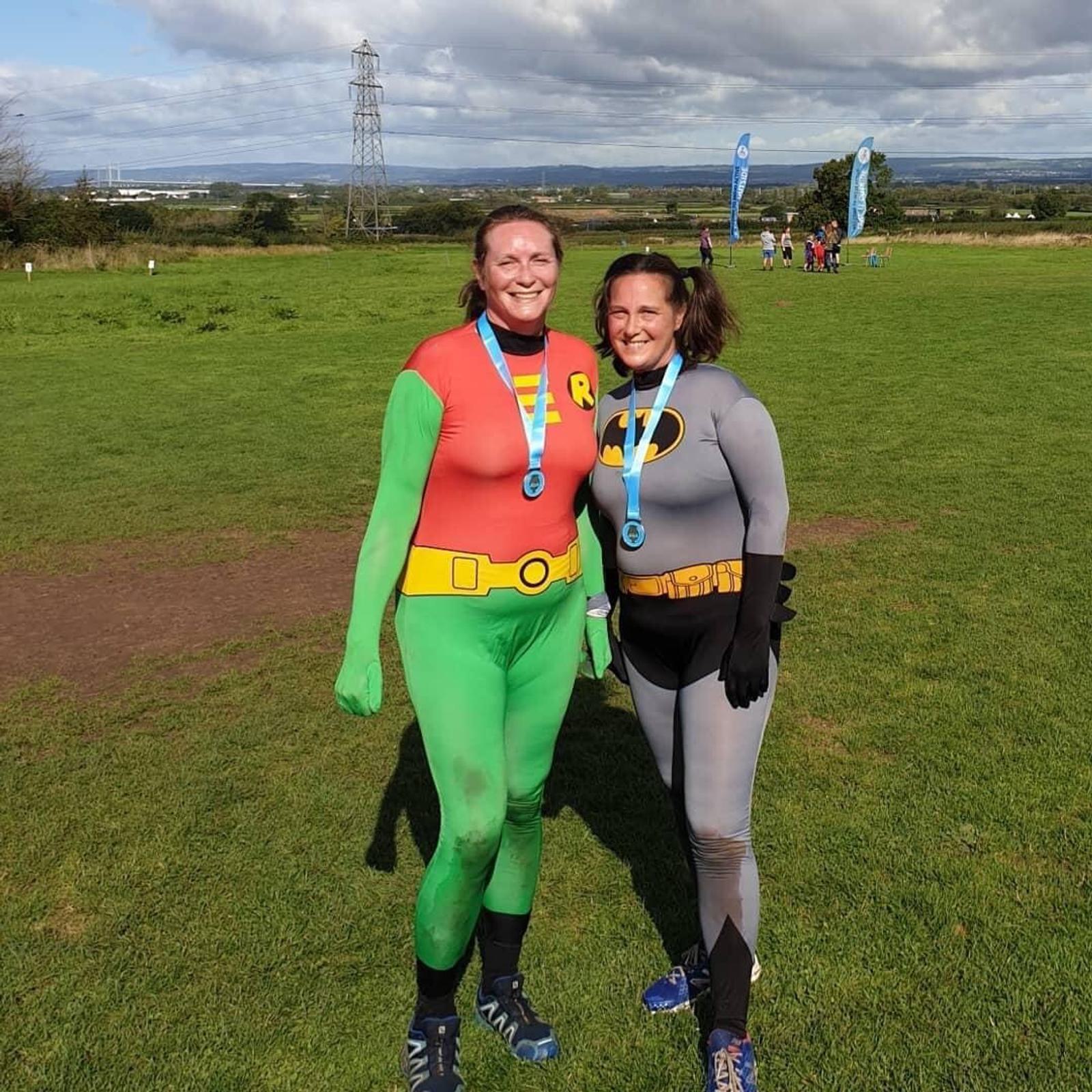 Two adults dressed as Robin and Batman stand on a grassy field with medals around their necks, smiling at the camera. Other individuals and event banners are visible in the background under a partly cloudy sky.
